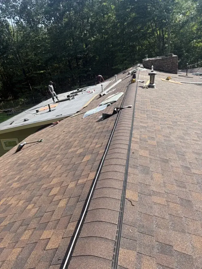 New ridge vent and flashing installed along a shingle roofline, with a flat roof section and stone chimney in the background